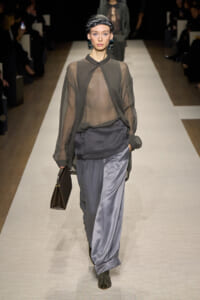 Model walking a runway in a sheer gray blouse over a draped top, paired with satin gray wide-leg pants, dark headscarf, bold green earrings, black boots, and a brown handbag.