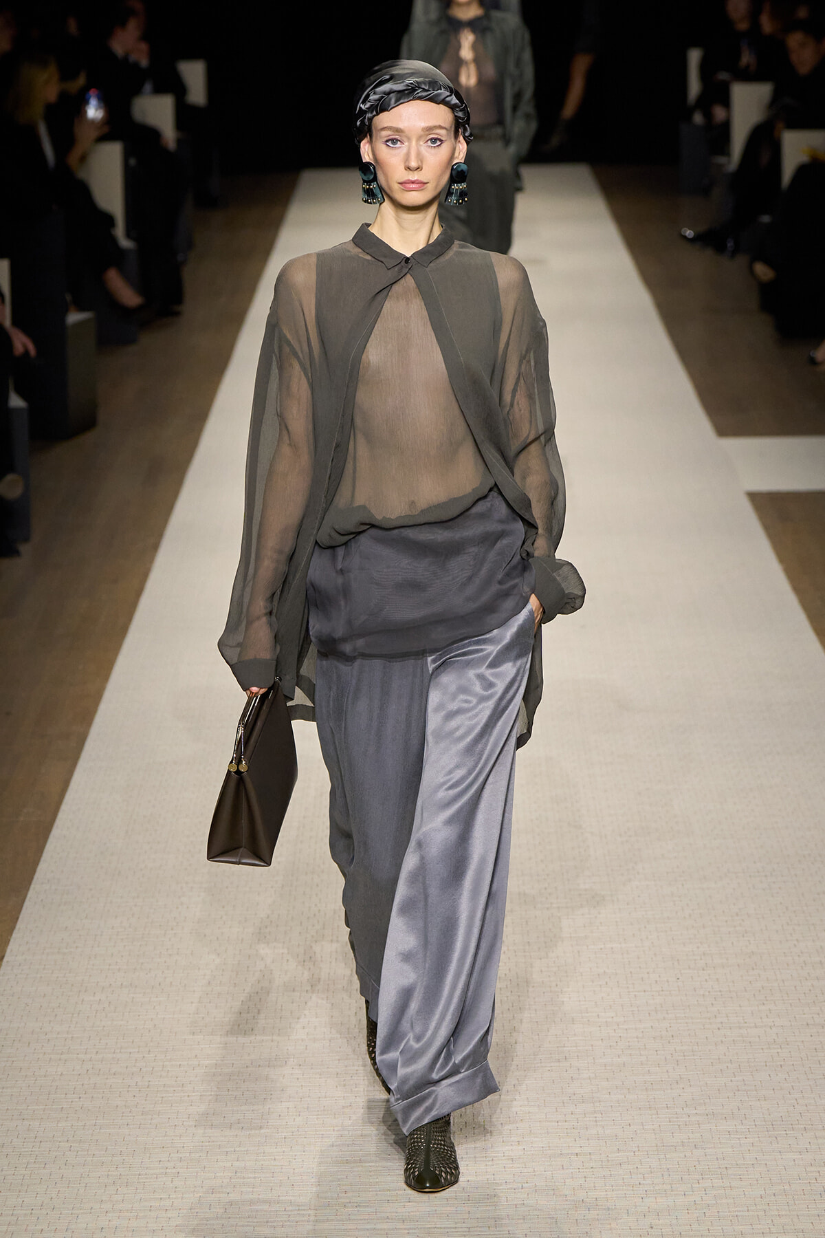 Model walking a runway in a sheer gray blouse over a draped top, paired with satin gray wide-leg pants, dark headscarf, bold green earrings, black boots, and a brown handbag.