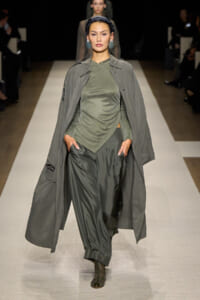 Model walking a runway in an olive-green wrap top, wide olive pants, and a long gray coat draped over the shoulders, with teal earrings and a headband.