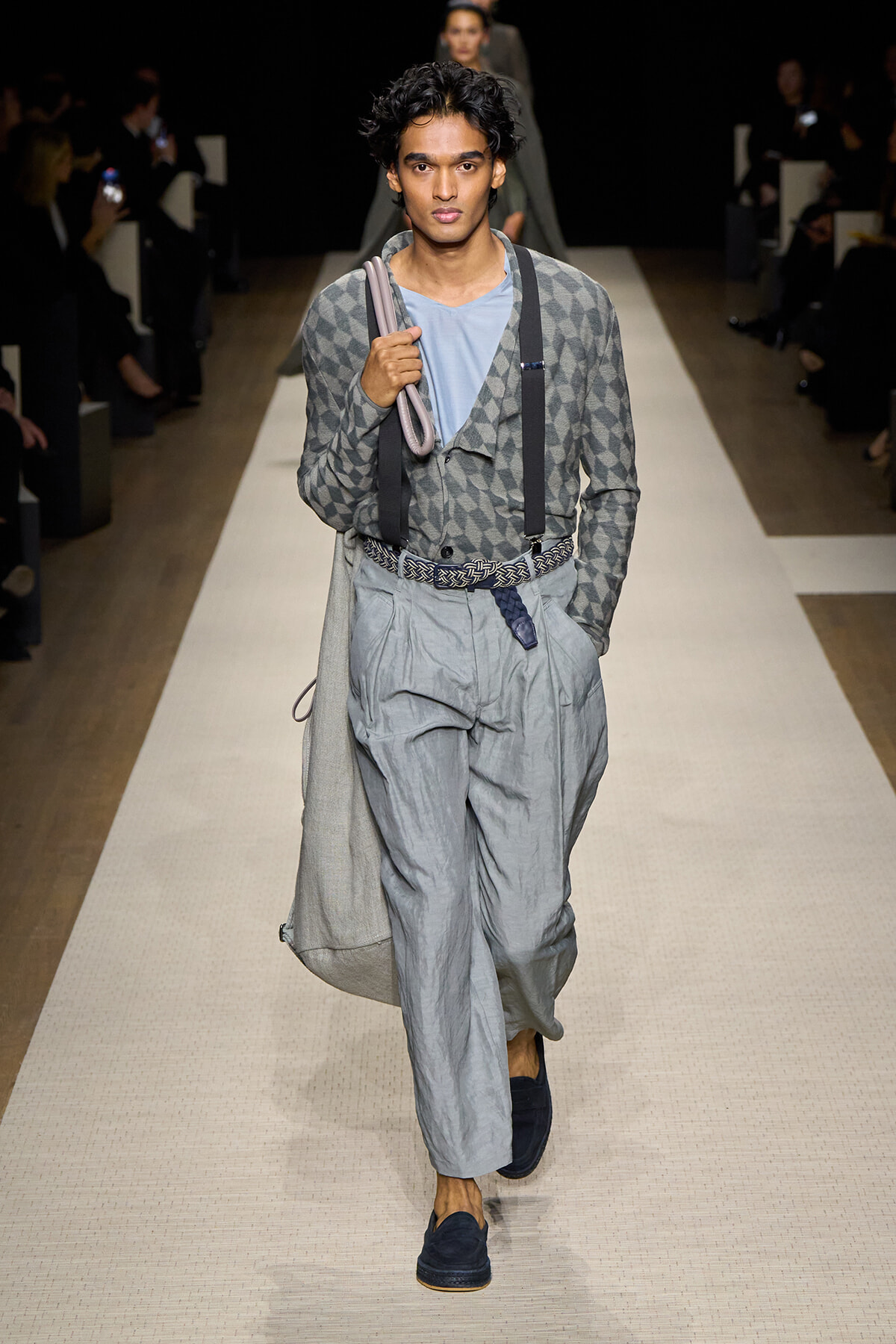 Male model walks the runway in a gray geometric-pattern cardigan, light blue shirt, suspenders, and wide gray trousers, with a long coat draped over his shoulder.