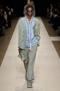 Male model walks a runway in a light teal suit with a pale blue shawl-collar shirt, sunglasses, and a woven bag in hand.