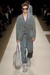 Male model walks a fashion runway wearing a gray-blue relaxed suit, sunglasses, and gray boots, carrying a light gray tote bag.
