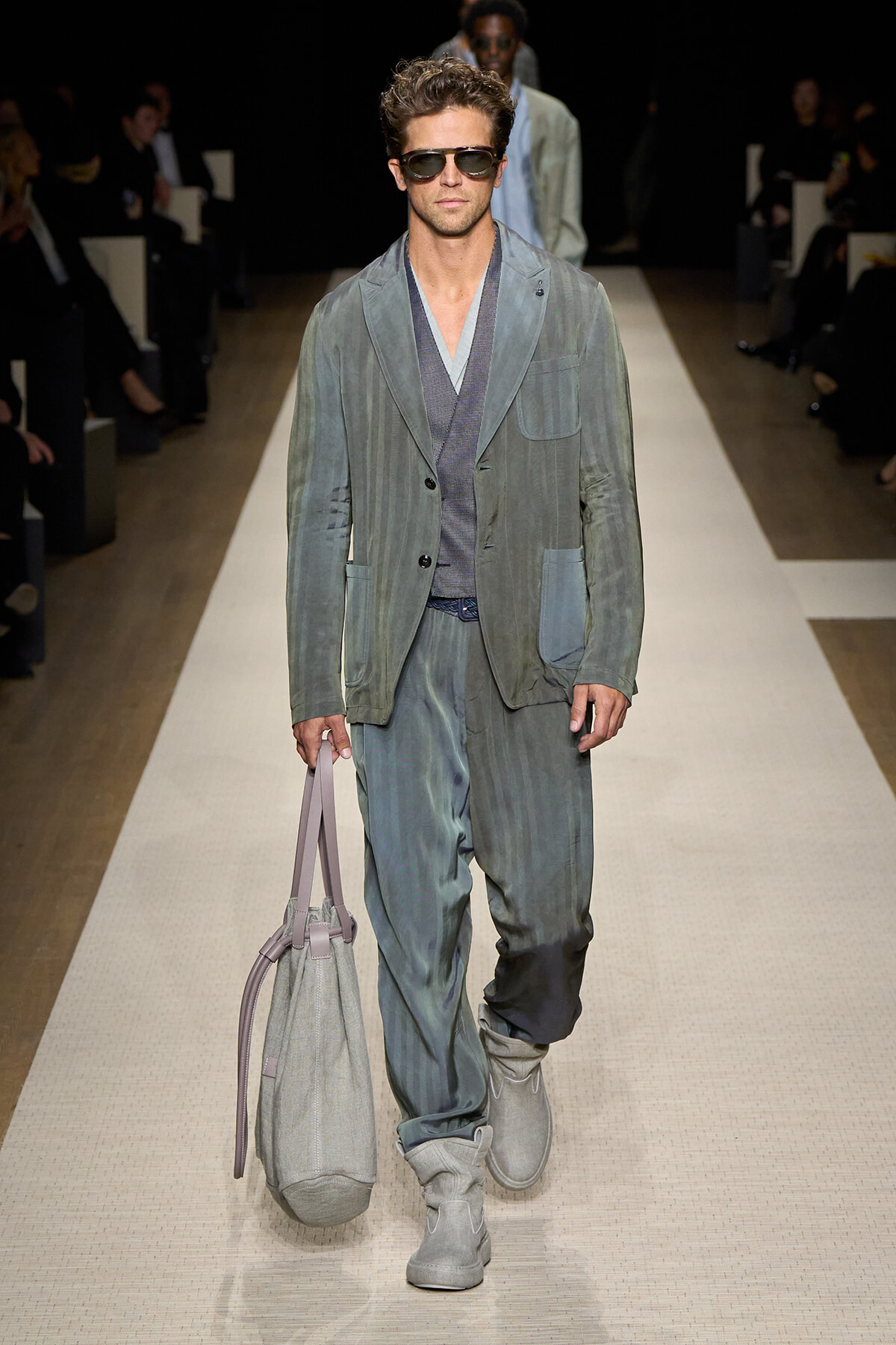 Male model walks a fashion runway wearing a gray-blue relaxed suit, sunglasses, and gray boots, carrying a light gray tote bag.