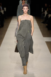 Model walking a runway in a loose, strapless grey draped jumpsuit with a brown handbag and tan cutout ankle boots.