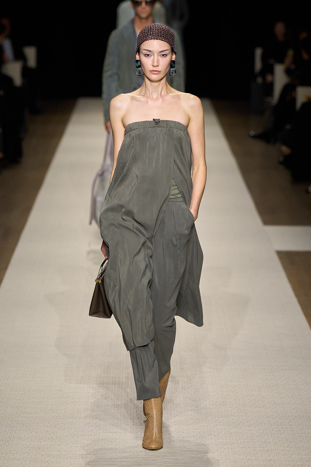 Model walking a runway in a loose, strapless grey draped jumpsuit with a brown handbag and tan cutout ankle boots.
