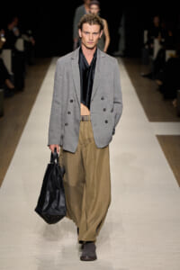 Male model on a runway wearing a light gray double-breasted blazer, black satin scarf, tan wide-leg trousers, and carrying a black leather bag.