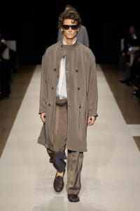 Male model walking a fashion runway in a loose taupe trench coat, light shirt, baggy trousers, and dark sunglasses.