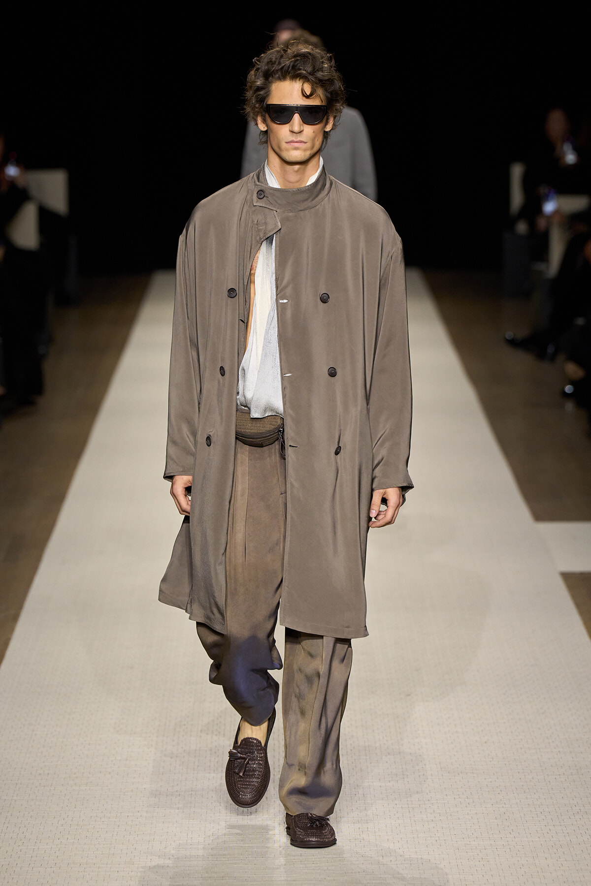 Male model walking a fashion runway in a loose taupe trench coat, light shirt, baggy trousers, and dark sunglasses.