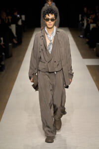 Male model walks a fashion runway in a loose taupe suit with draped vest, scarf, and sunglasses, holding a brown hat in one hand.