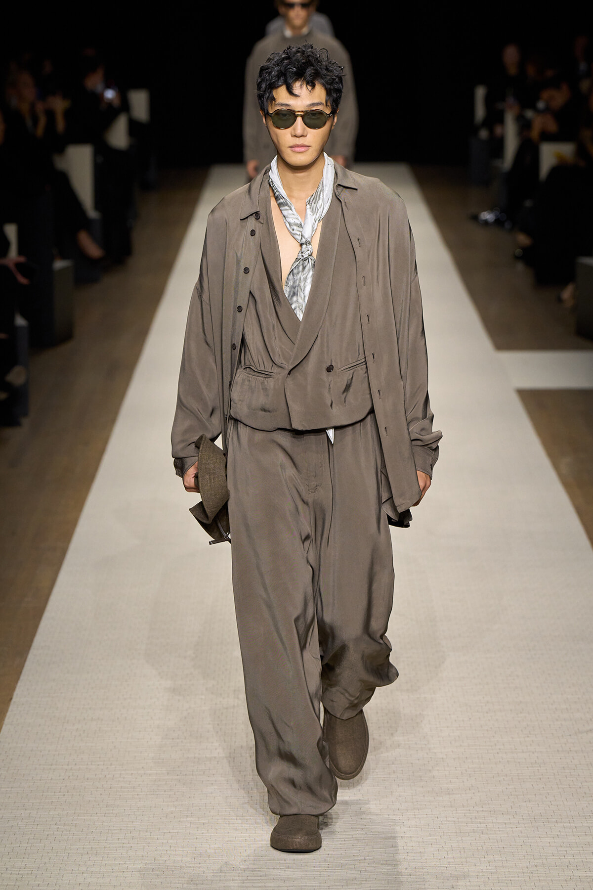 Male model walks a fashion runway in a loose taupe suit with draped vest, scarf, and sunglasses, holding a brown hat in one hand.