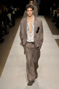 Male model walks a fashion runway in a taupe suit with a striped vest, open shirt, and sunglasses tucked in the jacket pocket.