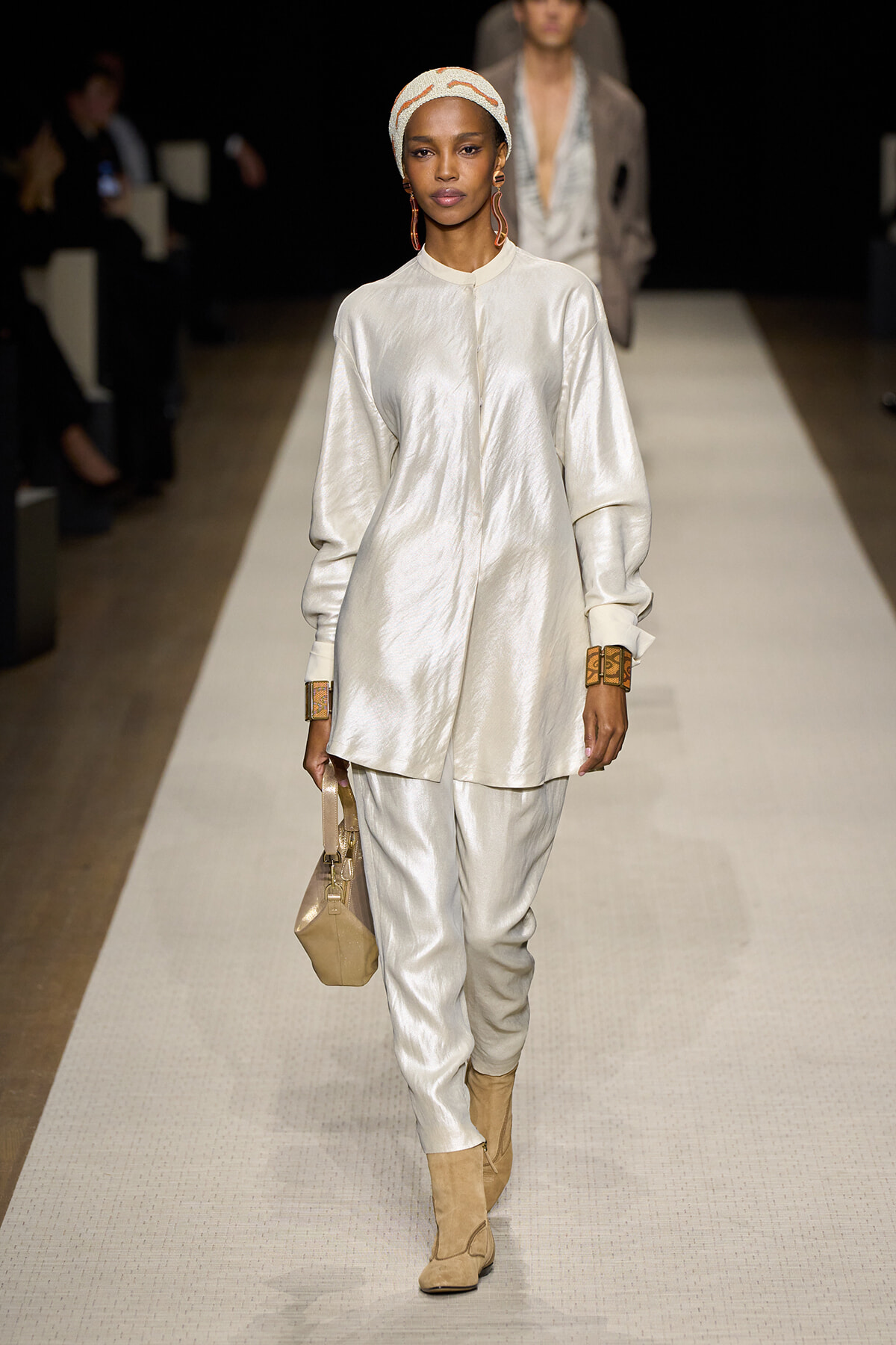 Model walking the runway in ivory satin oversized tunic and matching pants, beige headwrap, large earrings, tan suede ankle boots, and a beige handbag.