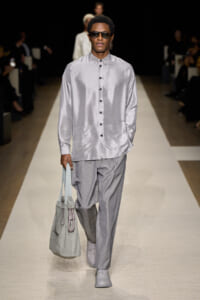 Male model walks a runway in a silvery satin button-down shirt and light gray trousers, carrying a gray tote bag and wearing sunglasses.