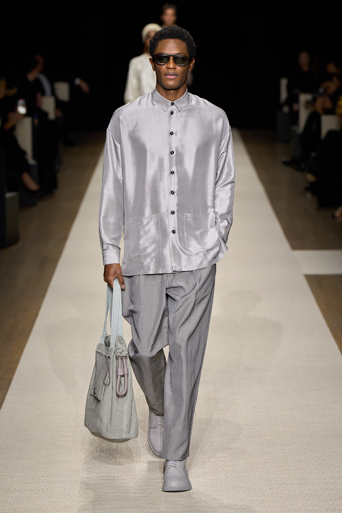 Male model walks a runway in a silvery satin button-down shirt and light gray trousers, carrying a gray tote bag and wearing sunglasses.