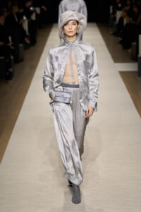 Model walks a fashion runway in a pale silver checkered suit with a sheer beige top, matching hat, gray belt bag, and gray ankle boots.