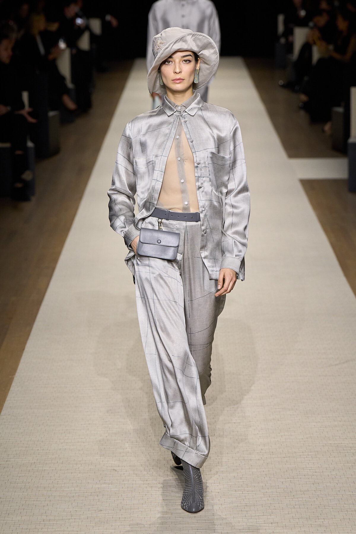 Model walks a fashion runway in a pale silver checkered suit with a sheer beige top, matching hat, gray belt bag, and gray ankle boots.