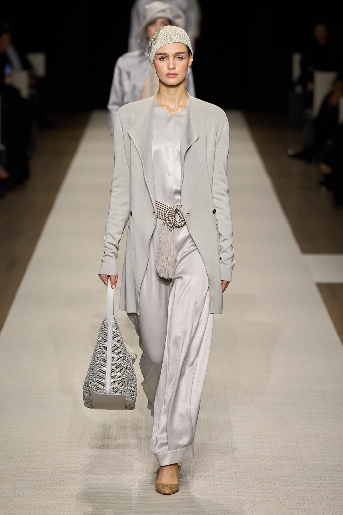 Model walks a runway in a monochrome gray ensemble: light gray blazer over a satin jumpsuit, braided belt with tassel, beige knit headwrap, and beige flats, carrying a lace-textured white tote bag while the audience watches in the background.