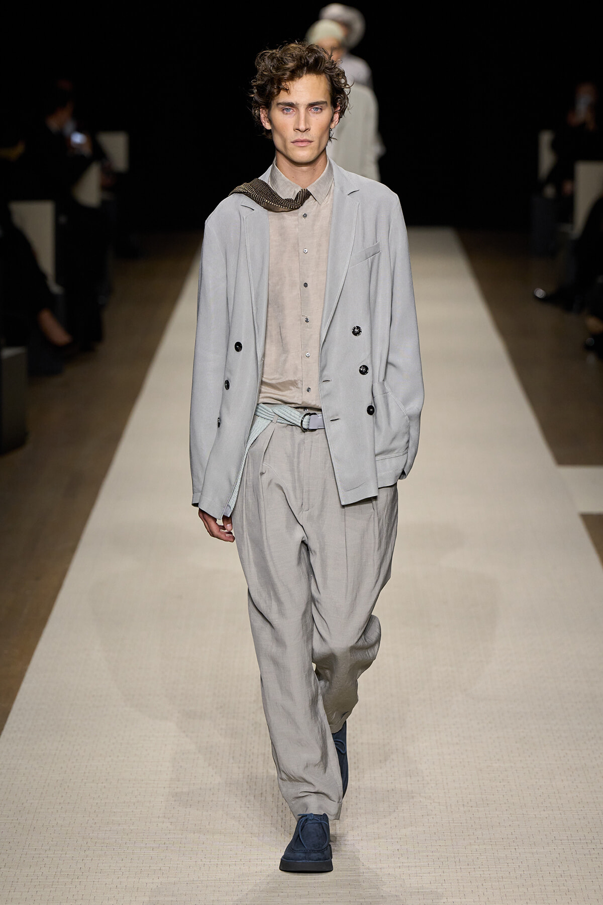 Male model walking a fashion runway in a light gray double-breasted suit with a beige shirt and blue shoes, neutral gaze.