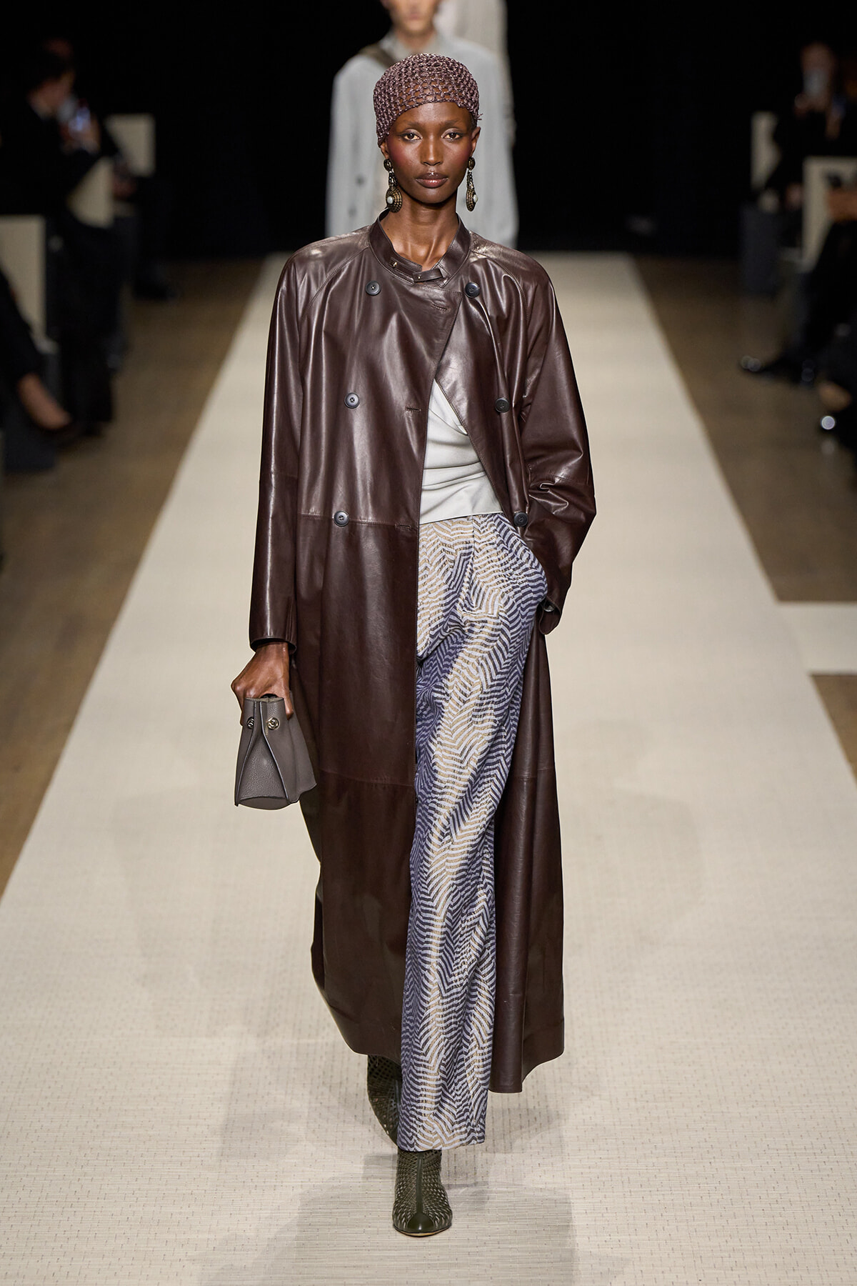 Model walks a runway in a long brown leather coat over a light top, patterned wide-leg pants, green ankle boots, and a beaded cap, carrying a gray handbag.