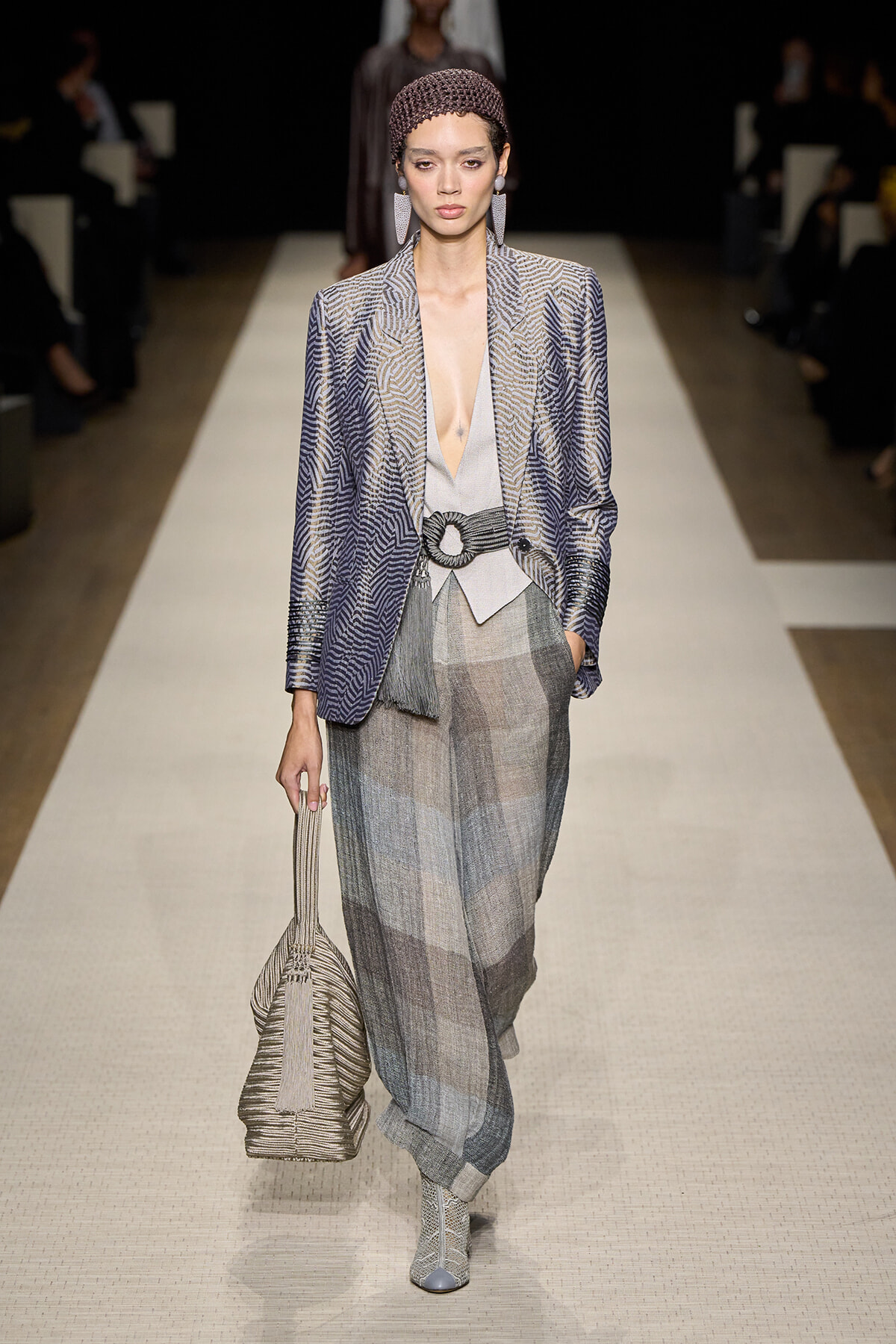 Model walks the runway in a metallic zigzag-pattern blazer over a plunging gray top, wide plaid trousers, and a large circular-buckle belt, paired with a knit headband and striped tote bag.