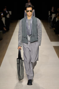 Male model walks a runway in a gray layered outfit: vest over a striped top, loose gray trousers, a metallic scarf, dark sunglasses, and a textured tote bag.