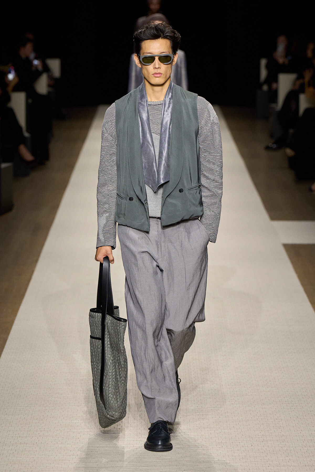 Male model walks a runway in a gray layered outfit: vest over a striped top, loose gray trousers, a metallic scarf, dark sunglasses, and a textured tote bag.