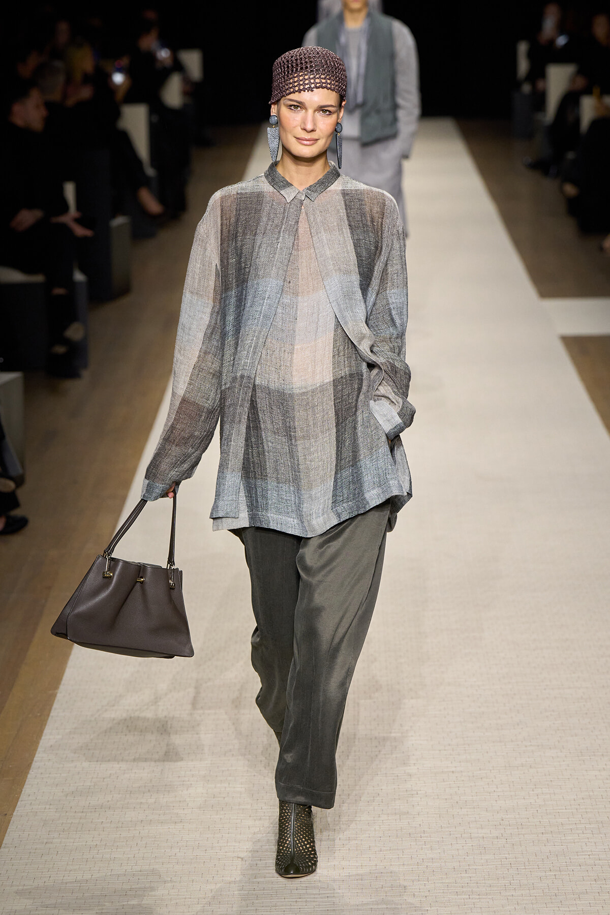 Model walks the runway in a loose gray plaid tunic over wide-leg pants, carrying a dark brown handbag.