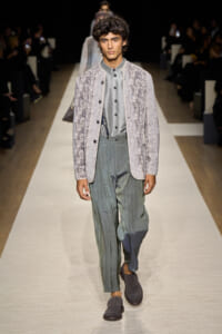 Male model walks a runway in a textured light-gray jacket over a gray button-up shirt and loose green-tinged pleated trousers, wearing gray loafers.