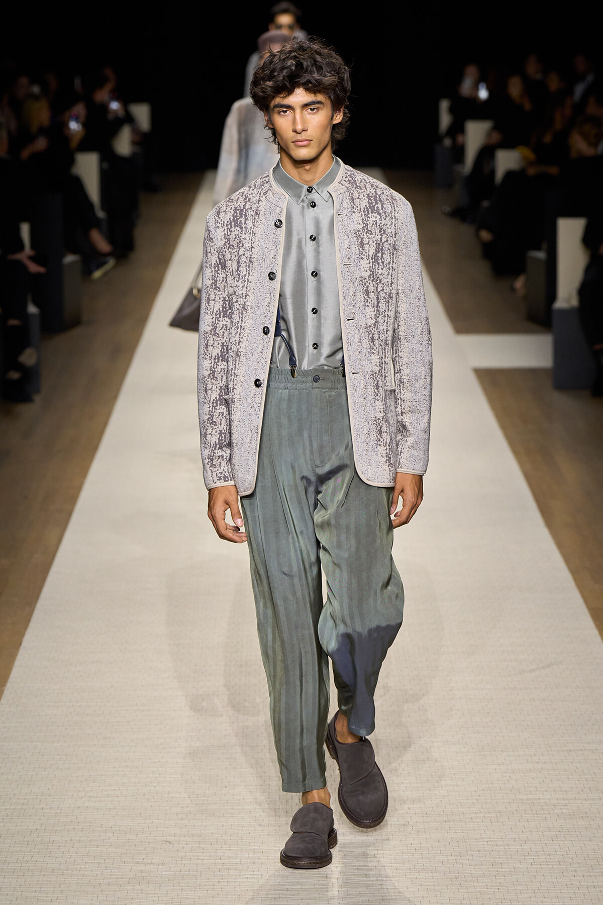 Male model walks a runway in a textured light-gray jacket over a gray button-up shirt and loose green-tinged pleated trousers, wearing gray loafers.