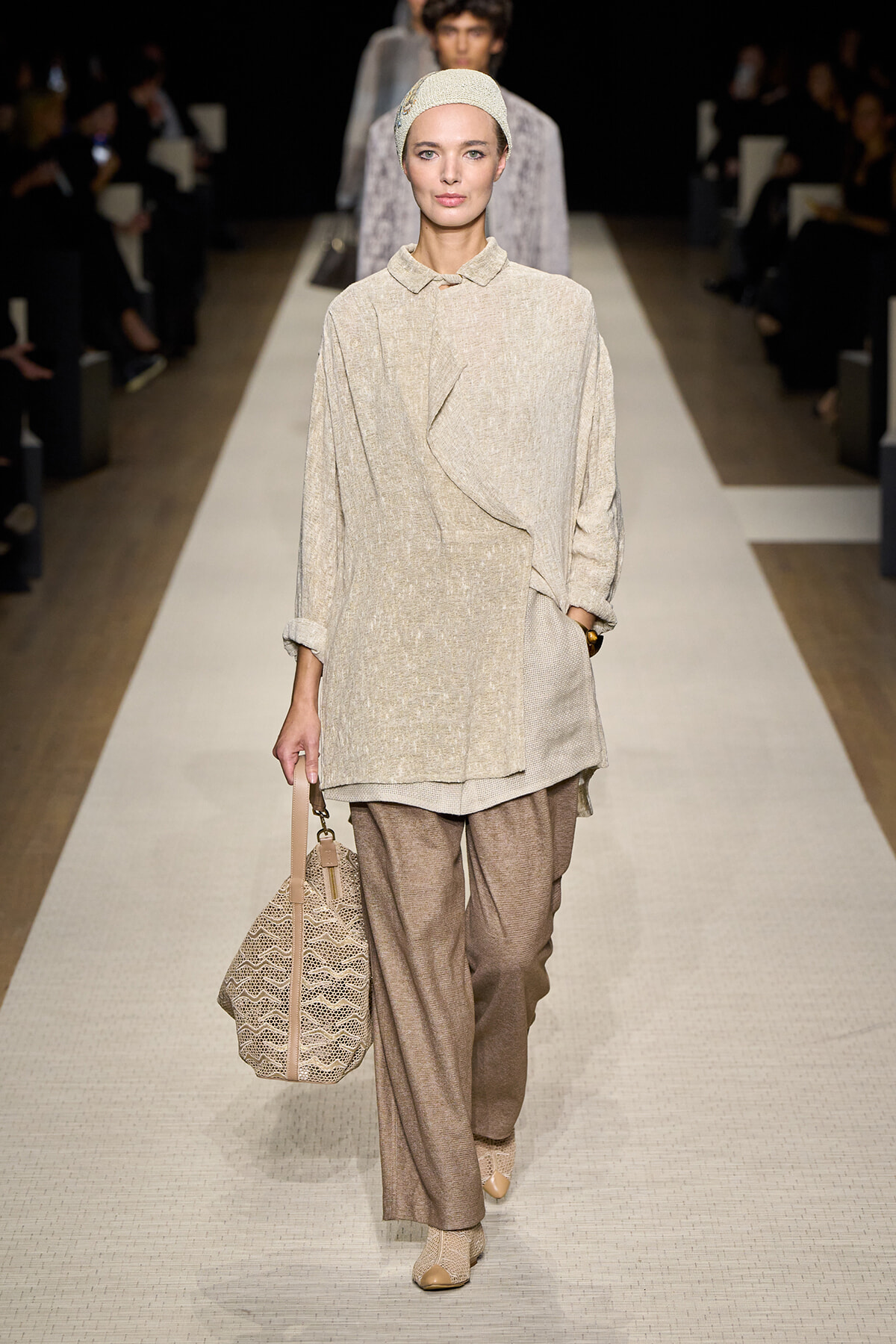 Model walks a neutral-toned runway wearing a beige textured blouse, loose pants, and a headwrap; carries a woven tote bag.