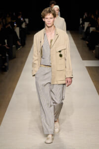 Male model walks a beige jacket over a light striped suit on a runway; sunglasses tucked in pocket.