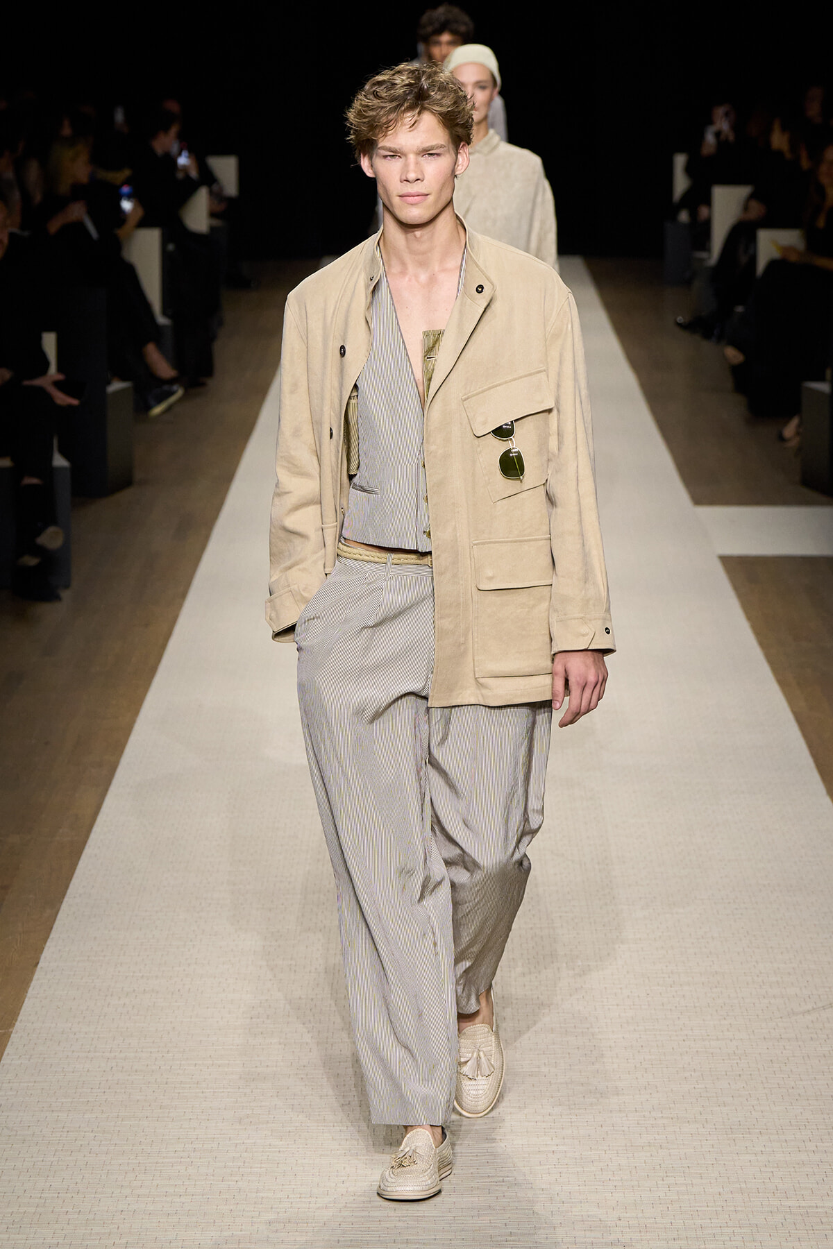Male model walks a beige jacket over a light striped suit on a runway; sunglasses tucked in pocket.