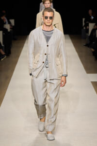 Male model walks the runway in a light beige linen suit with a striped shirt and sunglasses.