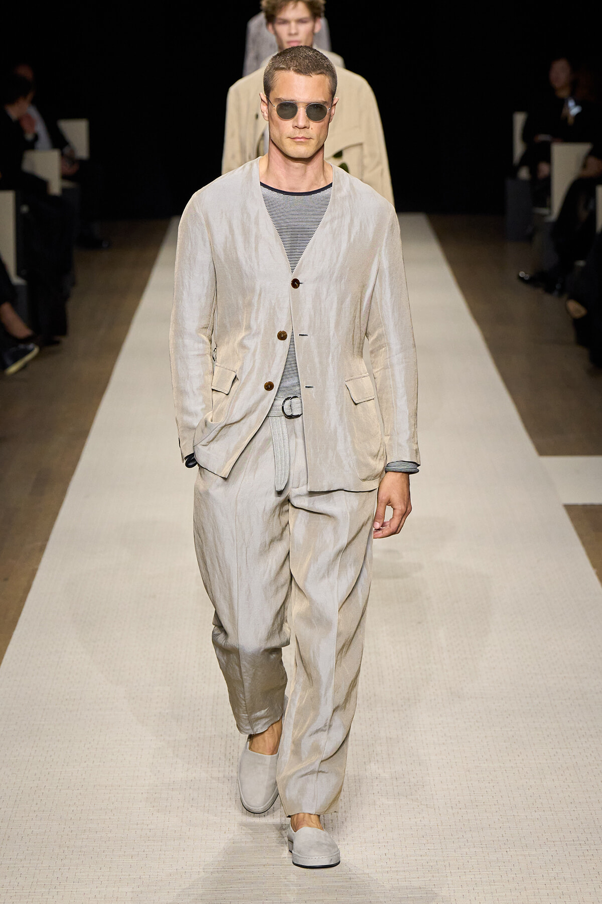 Male model walks the runway in a light beige linen suit with a striped shirt and sunglasses.