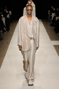 Male model walks the runway in a light beige textured suit with a gray top, patterned belt, and cream boots, wearing sunglasses.