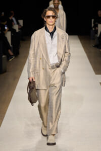 Male model walks a runway in a beige textured suit with a light shirt and black neck scarf, oversized sunglasses, and a brown bag.
