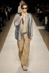 Male model on a runway wearing a metallic silver blazer, beige top, and wide tan pants, with sunglasses and a brown shoulder bag strap entering the frame.