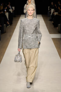 Model walking a runway in a gray knit tunic with a decorative white belt, beige wide-leg trousers, and a metallic headwrap, carrying a silver handbag and wearing patterned ankle boots.