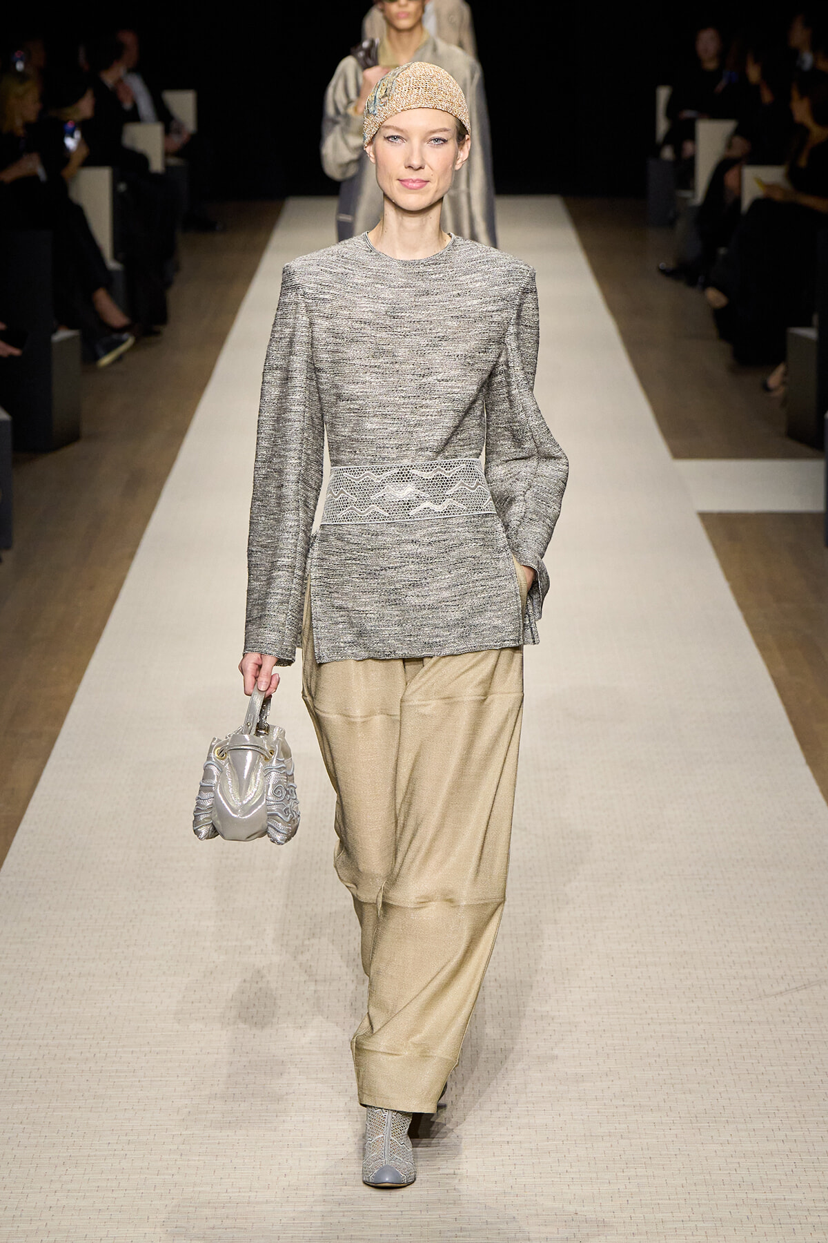 Model walking a runway in a gray knit tunic with a decorative white belt, beige wide-leg trousers, and a metallic headwrap, carrying a silver handbag and wearing patterned ankle boots.