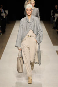 Model walks a runway in a light beige suit with a loose gray draped cardigan, matching head wrap, and beige tote bag.