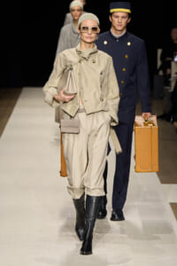 Model in a beige trench and matching pants walks the runway with beige handbag and clutch, wearing large sunglasses and black boots; a second model in a navy uniform follows with a tan suitcase behind her.