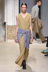 Model walks the runway in a gold sleeveless V-neck dress with a light blue knit sweater tied at the waist and black ankle boots.