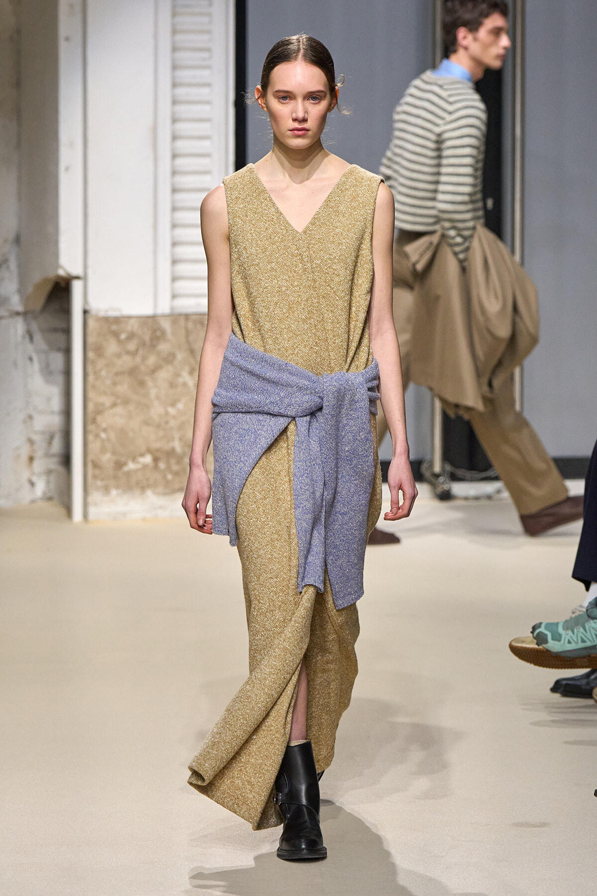 Model walks the runway in a gold sleeveless V-neck dress with a light blue knit sweater tied at the waist and black ankle boots.
