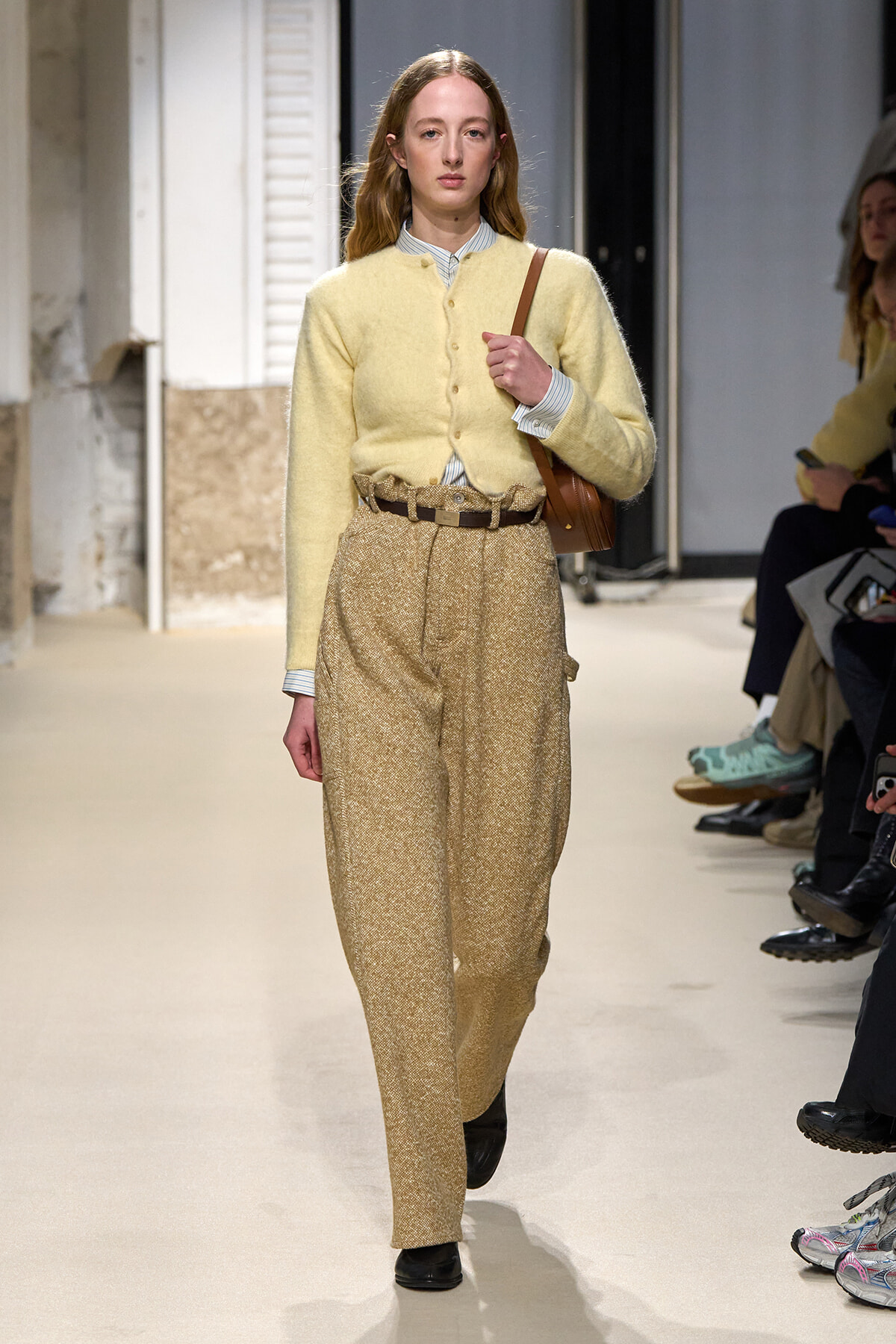 Model walking a fashion runway in a pale yellow cardigan, striped shirt, and high-waisted beige glitter trousers with a brown shoulder bag.