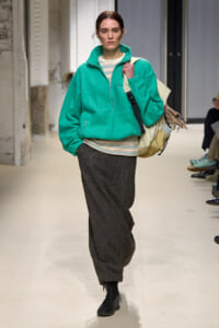 Model on a runway wearing a teal oversized fleece jacket over a striped top, brown wide-leg trousers, black boots, and a beige bag.
