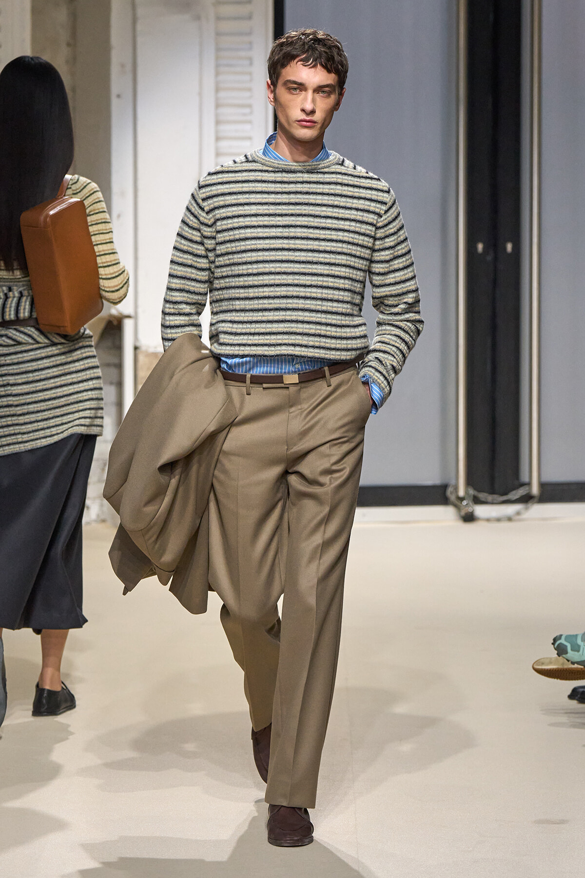 Male model walking a runway in a striped knit sweater, blue shirt, and tan wide-leg trousers, carrying a beige coat.