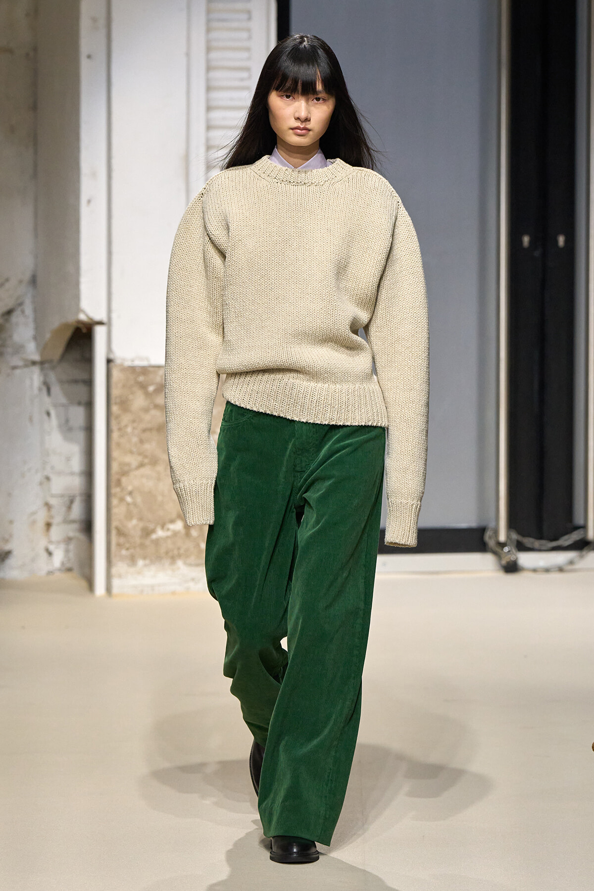 Model walking runway in a chunky beige knit sweater and emerald green wide-leg pants, minimalist backstage setting behind.