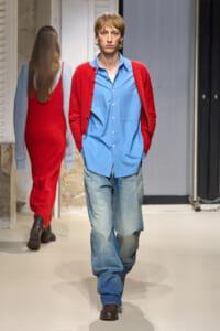 Male model walking a fashion runway in a light blue button-down shirt, red cardigan, and loose jeans with brown boots.