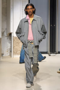 Male model walks a fashion runway in a loose gray bomber-style jacket over a pink sweater, pale yellow top peeking, with a striped belt tied around the waist.