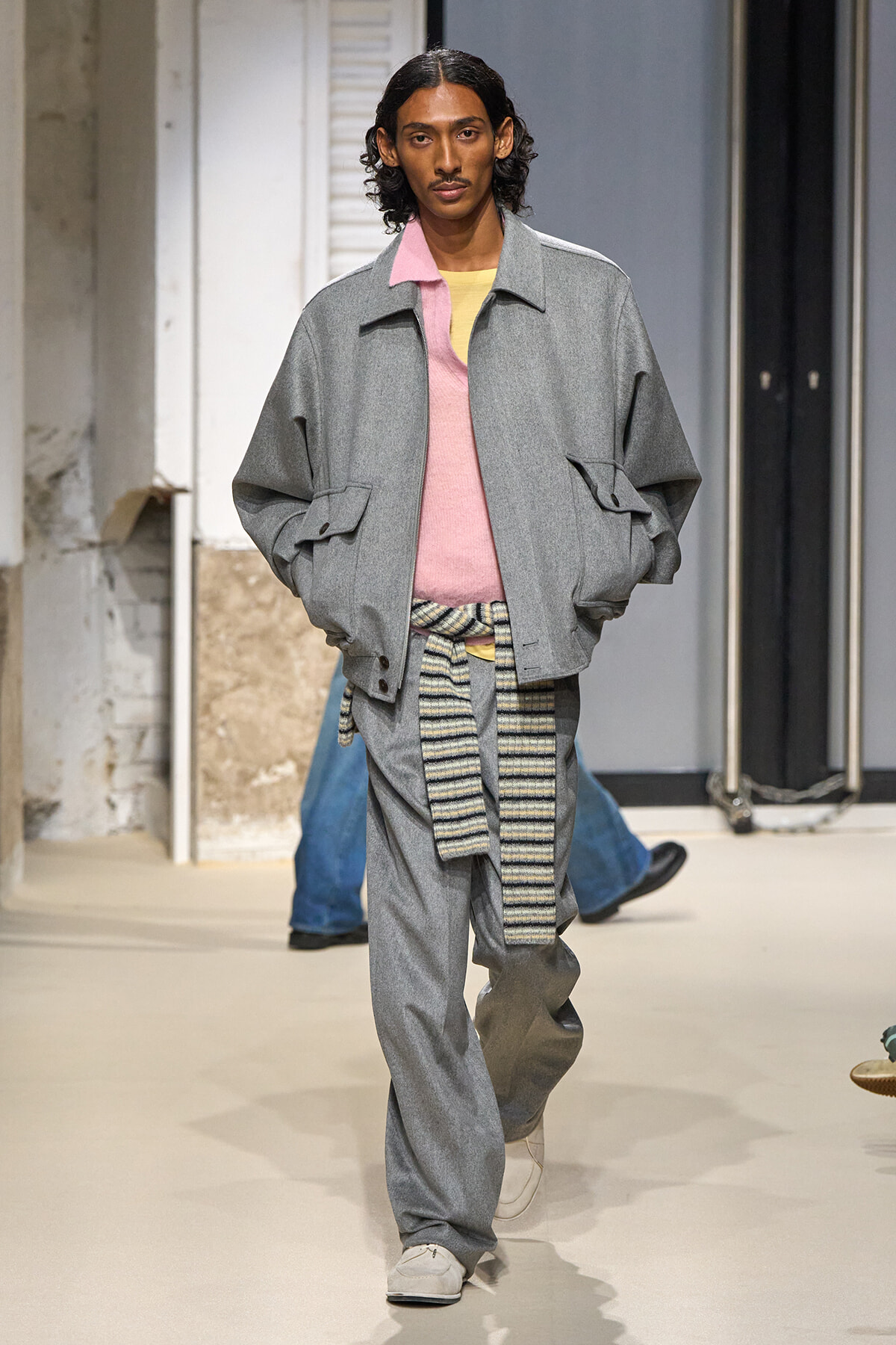Male model walks a fashion runway in a loose gray bomber-style jacket over a pink sweater, pale yellow top peeking, with a striped belt tied around the waist.